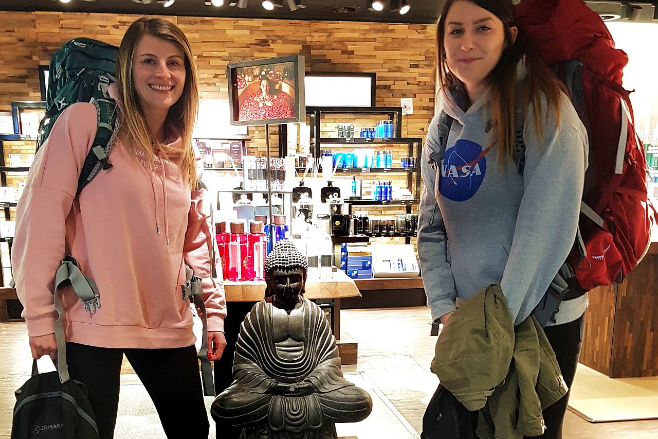 Zwei Backpackerfrauen mit Rucksack am Flughafen vor einer Buddhastatue